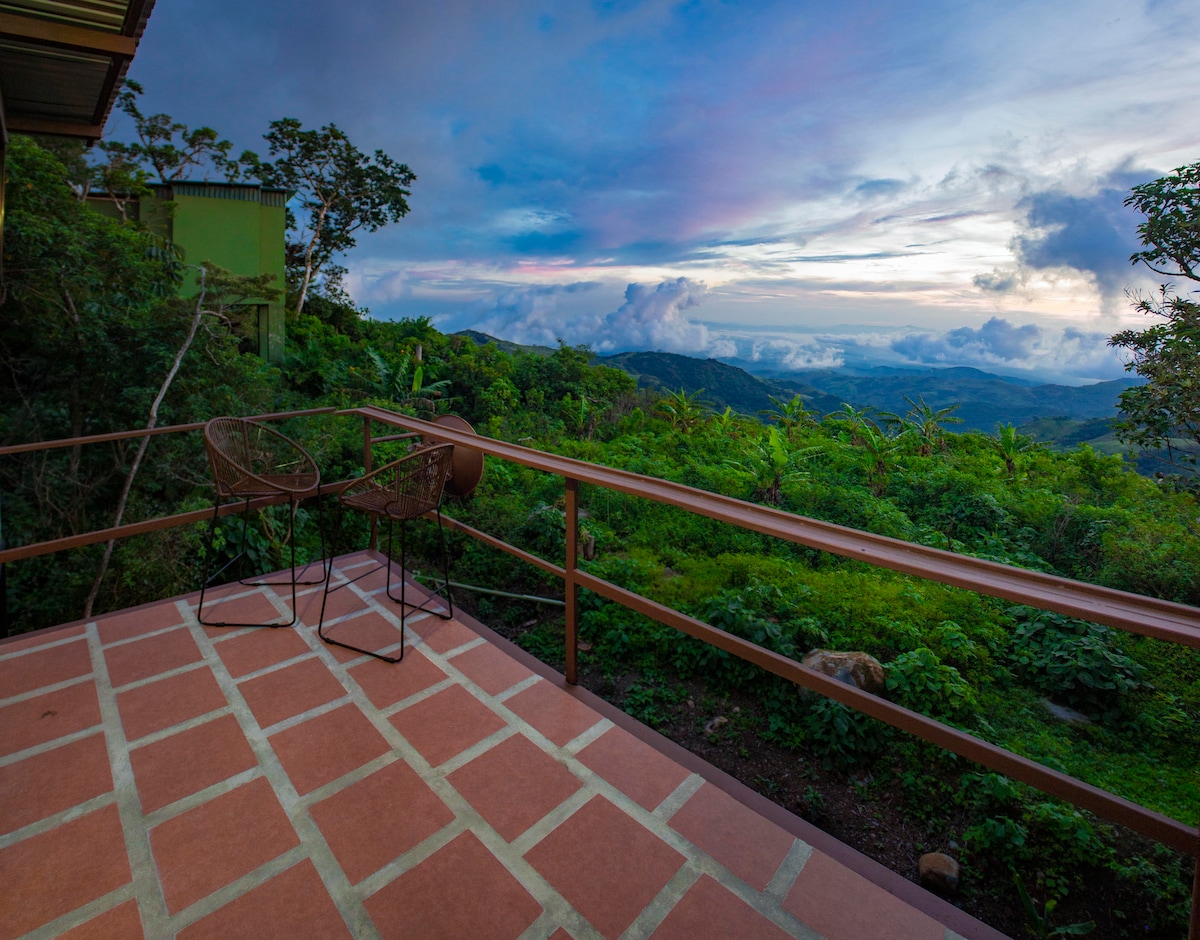 A covered balcony is seen with two high stools positioned at a small table. The floor features a tiled pattern, and the lush green landscape stretches into the distance under a colorful sky at twilight. Trees and plants surround the area, enhancing the view.