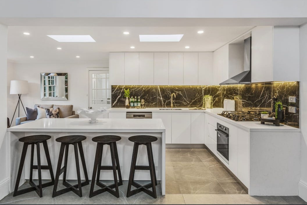 A modern kitchen is showcased with sleek white cabinetry and dark marble accents. A central island offers space for four stools. Skylights illuminate the area, featuring essential appliances and utensils neatly arranged, contributing to a functional cooking environment.