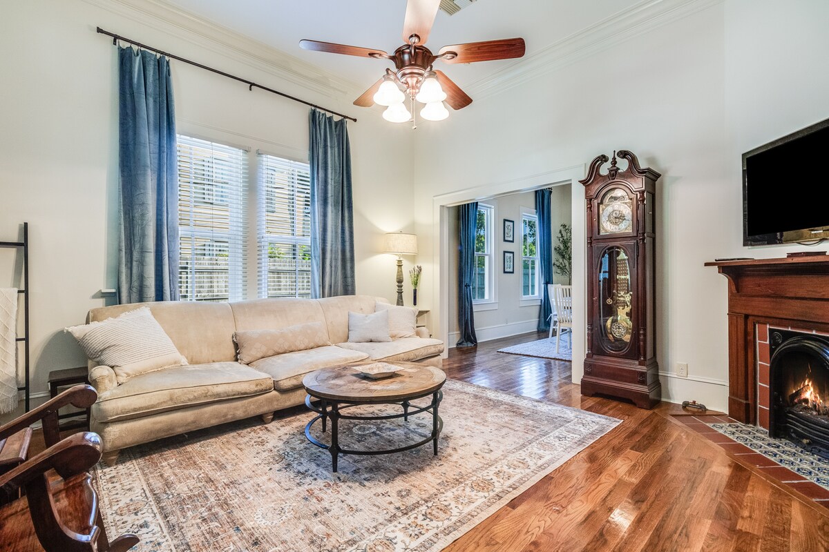 A spacious living area is highlighted by a light-colored sofa and decorative area rug on polished hardwood floors. Natural light filters through large windows adorned with blue curtains, while an antique clock stands prominently beside a small television and a cozy fireplace.