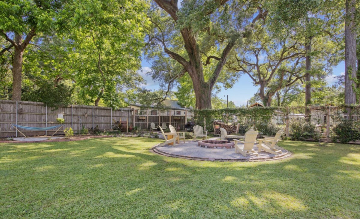 A spacious backyard is framed by lush greenery and large oak trees. A circular fire pit area with several light-colored Adirondack chairs is visible on well-maintained grass. A hammock can be seen in the background, inviting relaxation in this serene outdoor space.