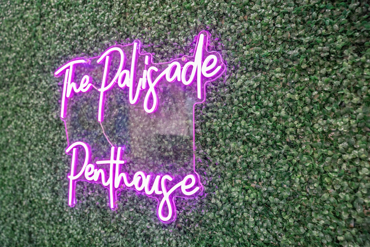A neon pink sign reading 'The Palisade Penthouse' is displayed against a textured green wall, evoking a vibrant yet inviting look. The sign contrasts with the greenery, providing a modern touch to the space.