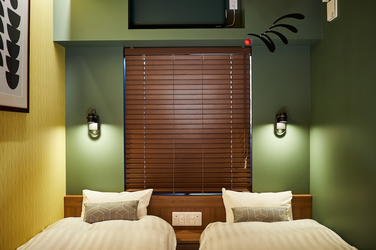The room features two spacious beds with white linens, positioned against green walls. Above, a flat-screen television is mounted. Soft lighting is provided by wall-mounted fixtures on either side of the beds, and wooden blinds cover the window for privacy.