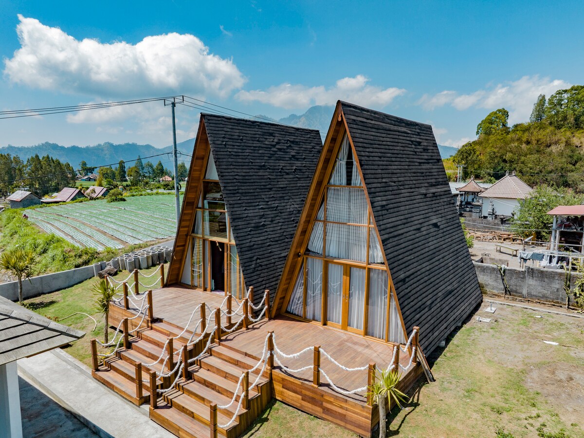 The chalet features a unique A-frame design with large glass windows, allowing for abundant natural light. A spacious wooden deck is accessible from the front, adorned with decorative ropes. Lush greenery and distant mountains create a serene backdrop for the welcoming structure.