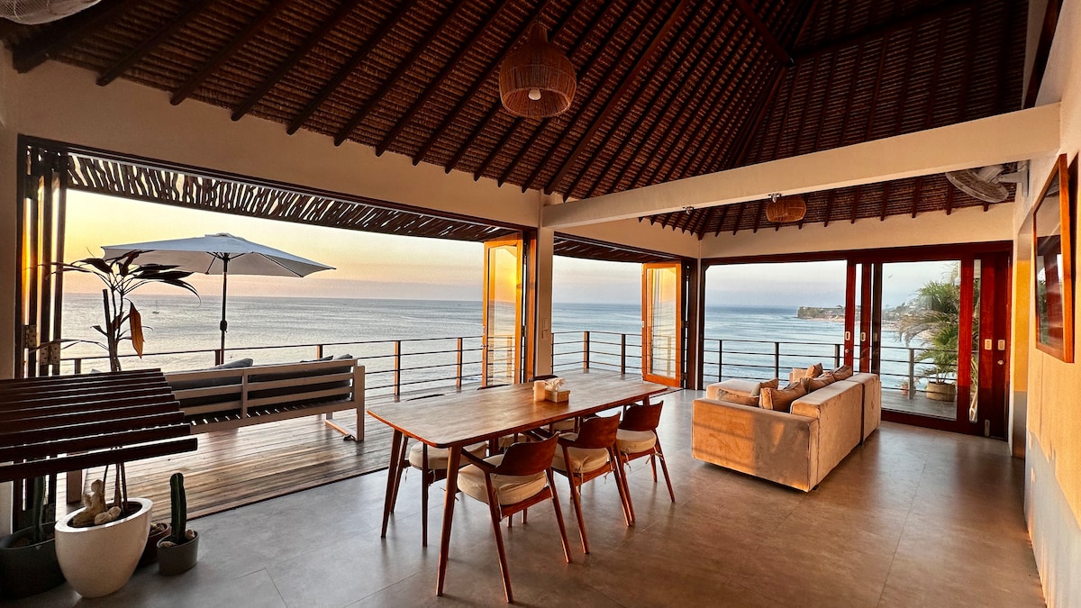 An open-plan living and dining area features large glass doors that provide unobstructed views of the ocean. A long dining table with wooden chairs centers the space, complemented by a spacious seating area. Natural light floods in as the sun begins to set.