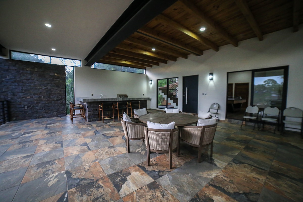 An open outdoor space features natural stone flooring and a rustic wooden ceiling. Seating is provided around a circular table, complemented by several additional chairs. A bar area with stools is positioned against a stone wall, enhancing the modern yet comfortable aesthetic.