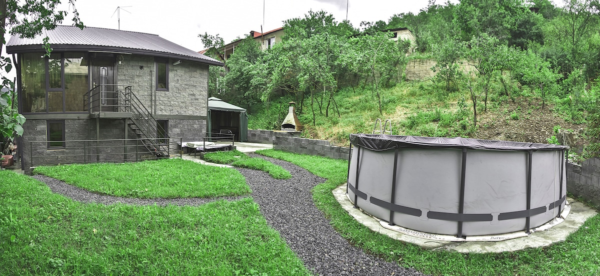 A modern house with a grey stone exterior is surrounded by lush greenery. A circular pool is positioned on a paved area, while a winding gravel pathway leads to the entrance. A charming outdoor fireplace can be seen off to the side.