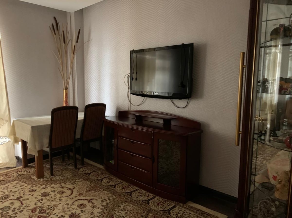 A living area is presented with a television mounted on the wall. A wooden sideboard is positioned beneath the TV, accompanied by a dining table surrounded by four chairs. A textured rug covers the floor, while decorative elements reach toward the ceiling.