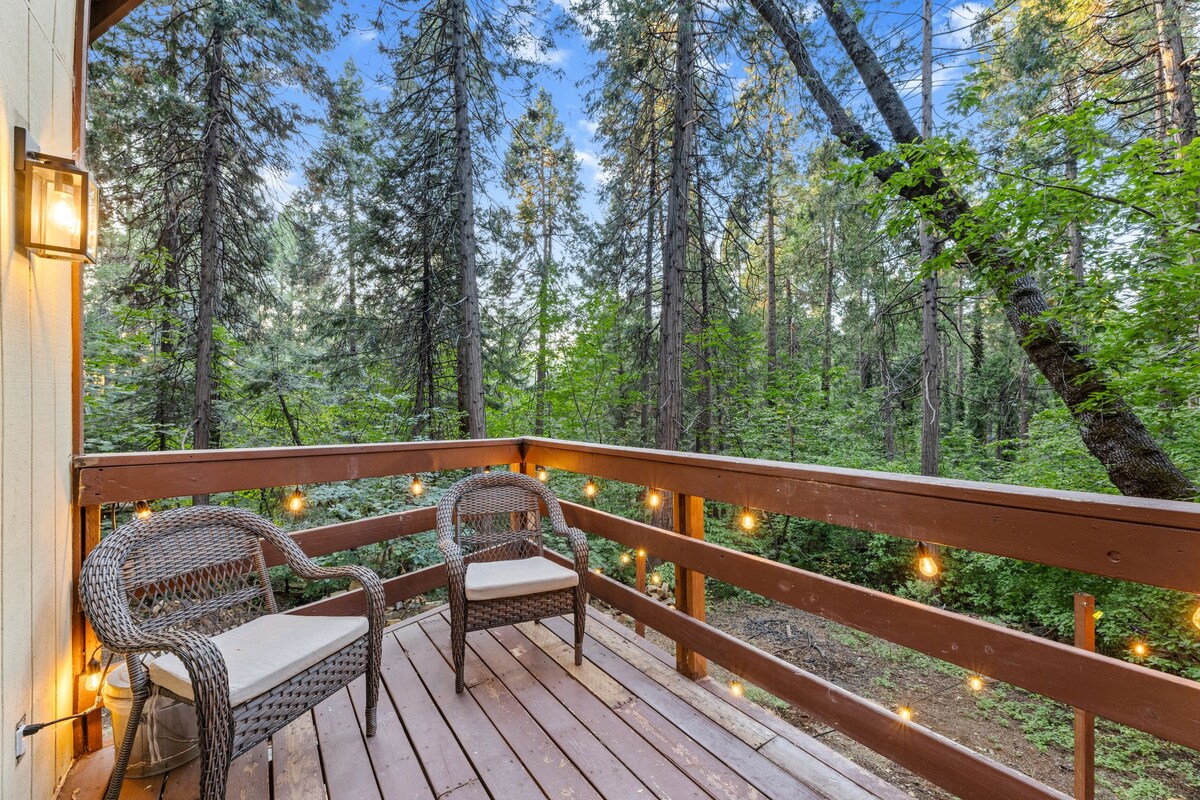A serene outdoor deck is surrounded by tall trees, featuring two woven chairs with light-colored cushions. Soft, warm string lights are hung along the railing, illuminating the space and enhancing the natural surroundings.
