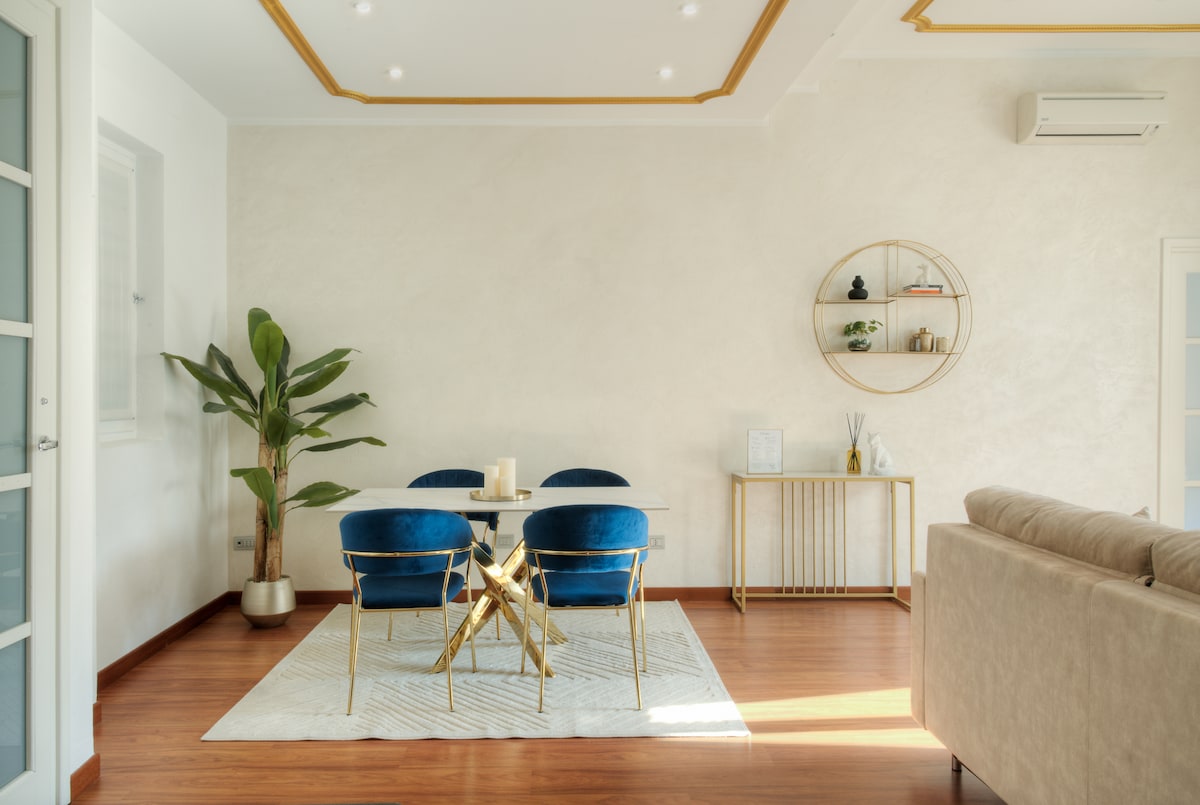 The dining area features a modern table surrounded by four blue velvet chairs. A small plant adds a touch of greenery. Behind the table, a decorative shelf displays minimalist decor. The room is well-lit, showcasing warm wooden flooring and subtle wall textures.