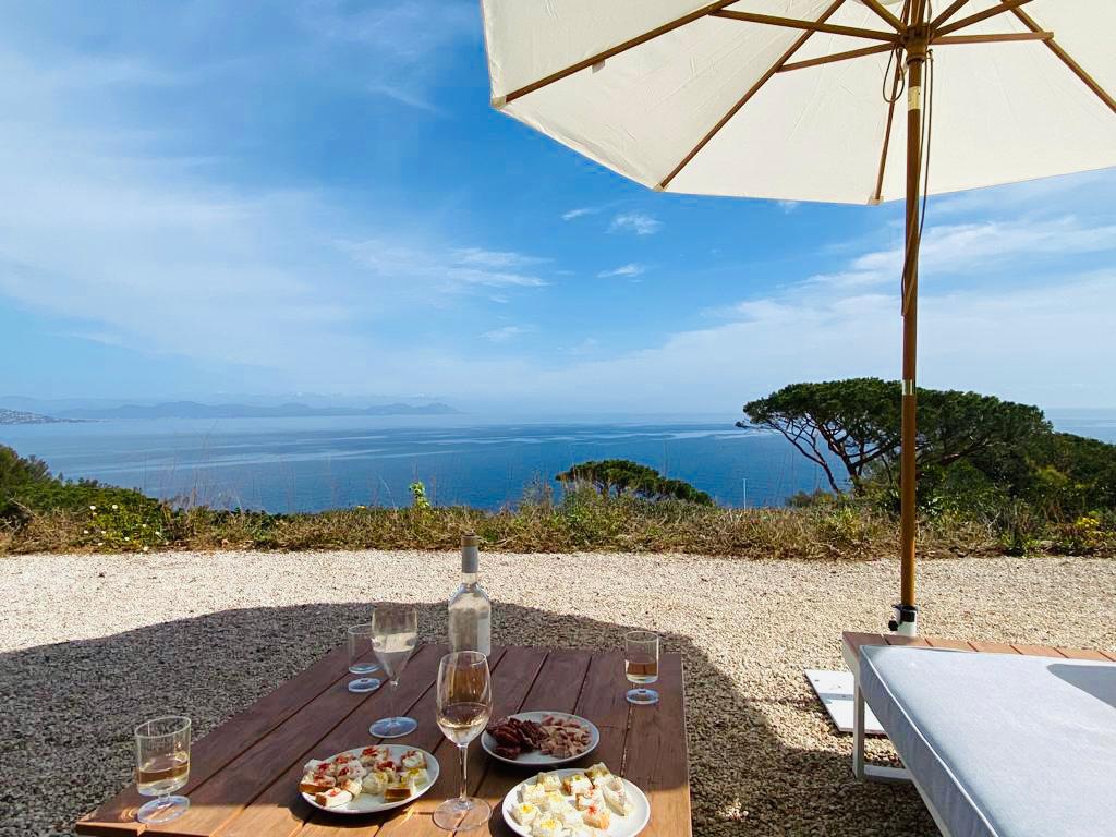 A wooden table is set with plates of food and glasses, situated beside a sunbed under a large umbrella. The tranquil sea stretches out in the background, with a clear sky above and distant land formations visible on the horizon.