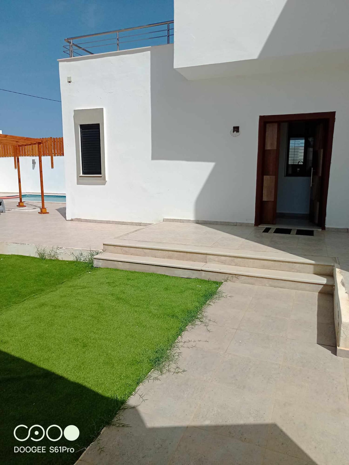 A well-maintained outdoor area is highlighted with manicured green grass and beige stone steps leading to an entrance. A bright, white exterior features large windows. A shaded section with wooden slats is visible in the background, providing a relaxing ambiance near the pool.