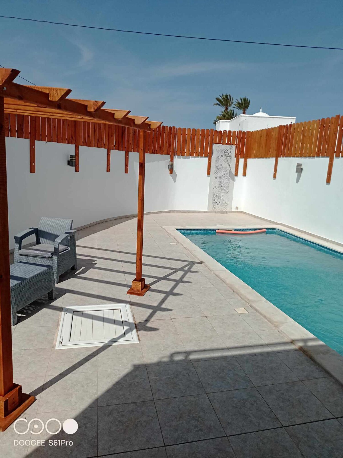 A tranquil pool area is featured, enclosed by a wooden pergola. The clear water of the pool reflects the sunshine, while gray lounge chairs provide seating. White walls and a tiled deck enhance the spacious feel, creating an inviting environment for relaxation.