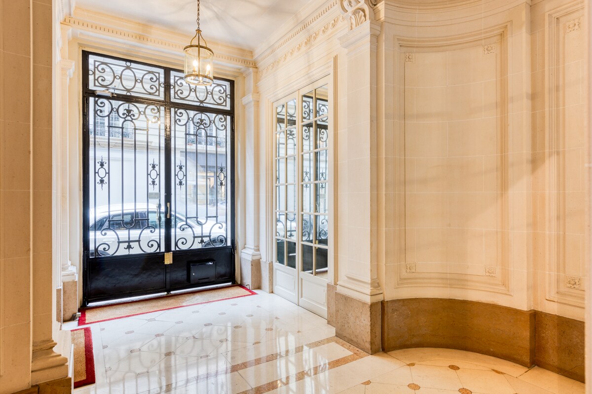 The elegant entrance hall features a mixture of polished marble flooring and classic panelled walls. A grand black double door with intricate ironwork allows natural light to fill the space, while a decorative light fixture hangs from the ceiling, enhancing the luxurious ambiance.