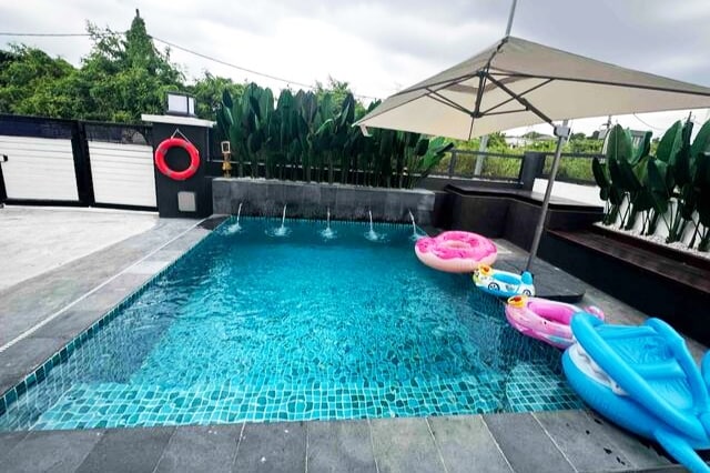 A refreshing swimming pool is featured, surrounded by a spacious stone deck. Colorful inflatable floats are present in the water, while an umbrella provides shade nearby. Lush greenery borders the area, enhancing the tranquil setting.