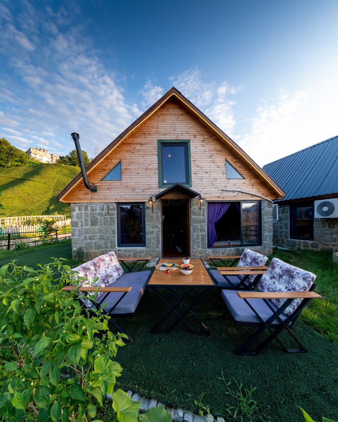 A charming bungalow is framed by a lush, green hillside. The exterior features a combination of stone and wood, and a spacious outdoor dining area with a wooden table is surrounded by four folding chairs. Large windows allow for natural light, enhancing the welcoming facade.