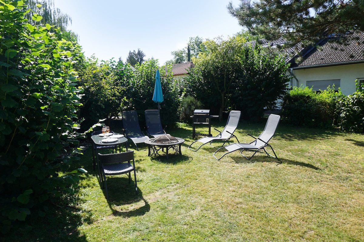 A lush garden area is featured, with several loungers arranged around a central fire pit. A dining table with chairs is present, shaded by an umbrella. Surrounding greenery offers a sense of privacy and tranquility.