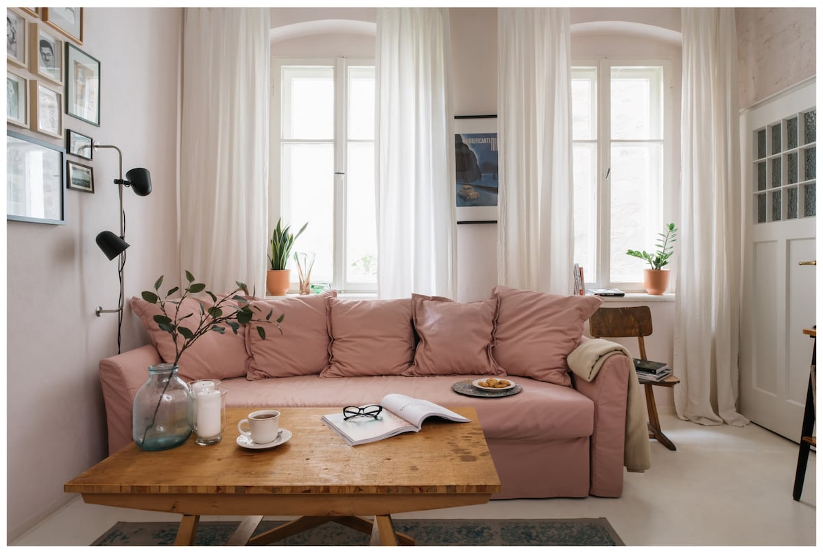 A light-filled living area features a spacious pink sofa with decorative cushions, positioned near large windows adorned with sheer curtains. A wooden coffee table holds a decorative glass bottle, a steaming cup, and an open book, while potted plants add a touch of greenery.