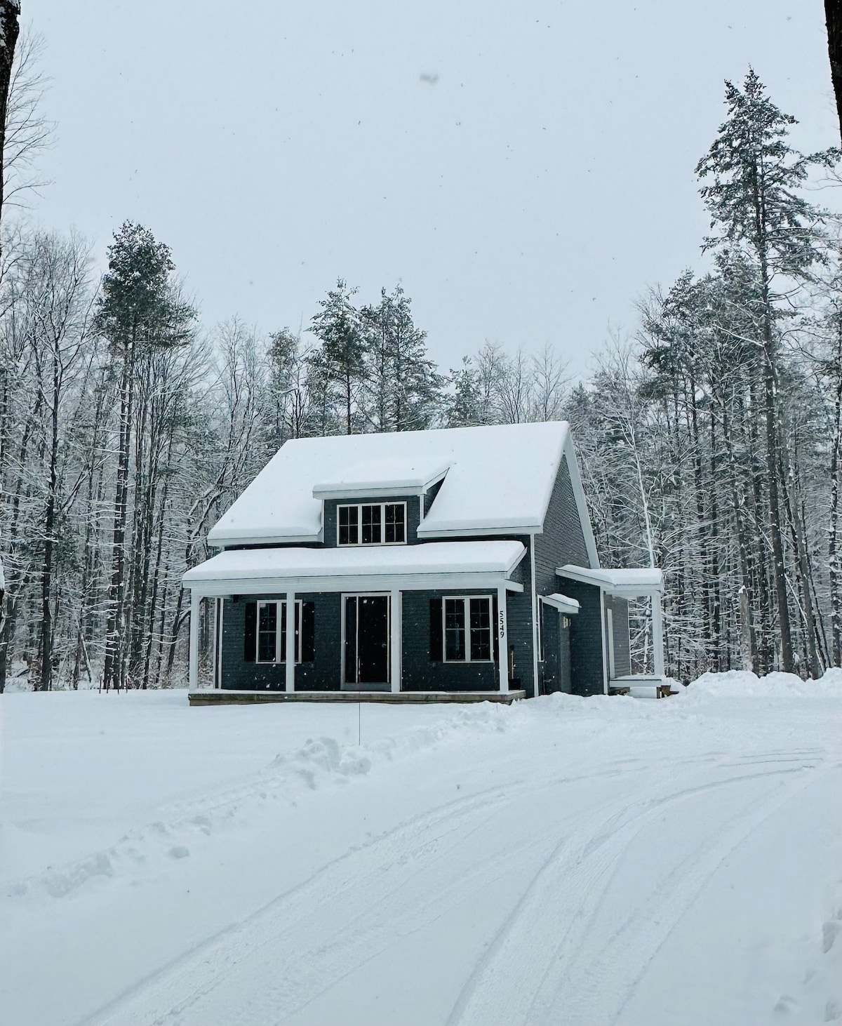 A new home is showcased amidst a snowy landscape, surrounded by towering trees. The structure features a covered front porch and prominent windows, allowing ample light to enter. Fresh snow blankets the ground, adding to the serene, wintery scene.