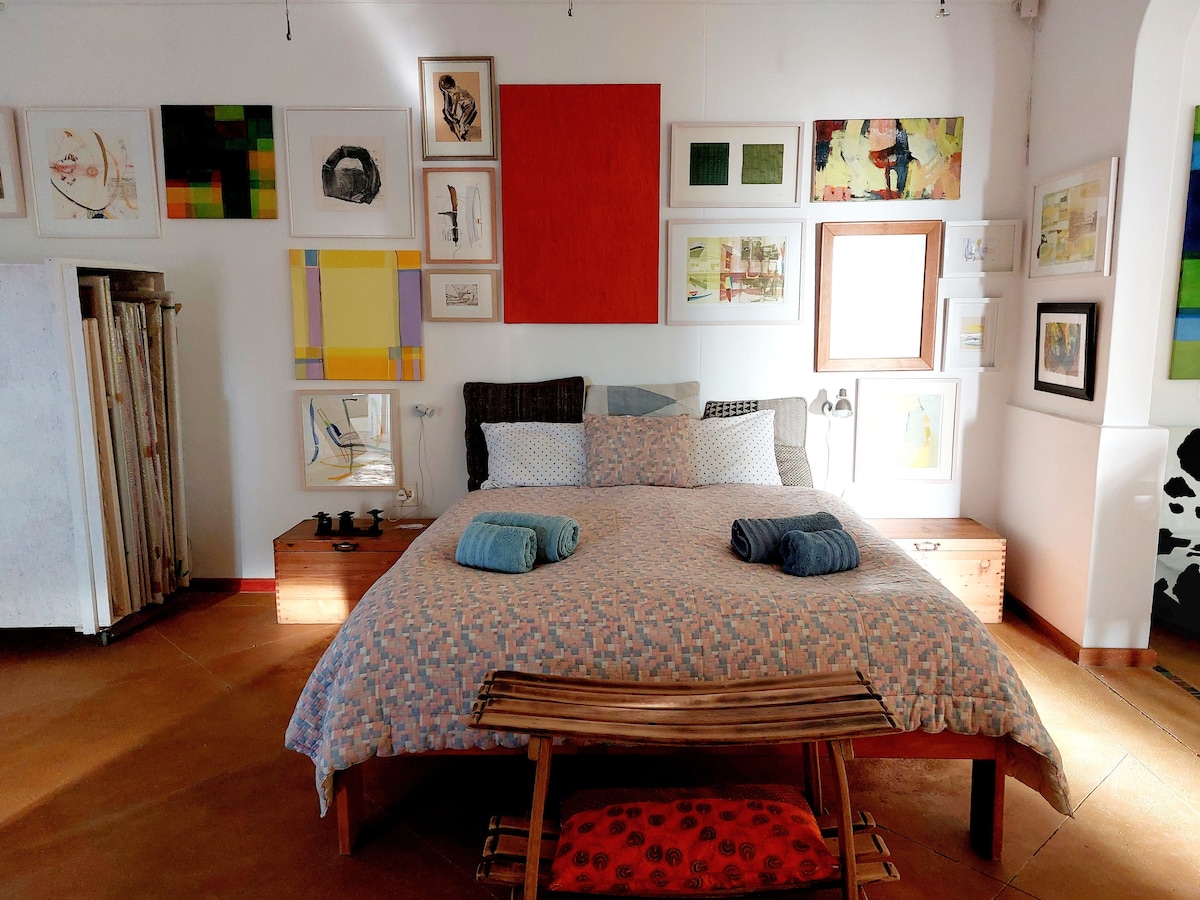 A spacious bed is centered in the room, adorned with a patterned duvet and flanked by two wooden nightstands. Colorful artwork in various frames decorates the walls. A bench sits at the foot of the bed, and two rolled towels are placed neatly on top.