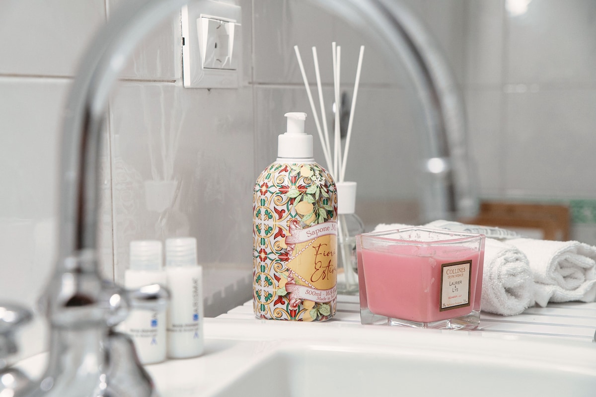 A clean bathroom sink is shown with decorative items. A stylish soap dispenser, adorned with a floral design, sits next to a pink candle in a glass holder. Nearby, neatly arranged toiletries and rolled towels add to the organized appearance.