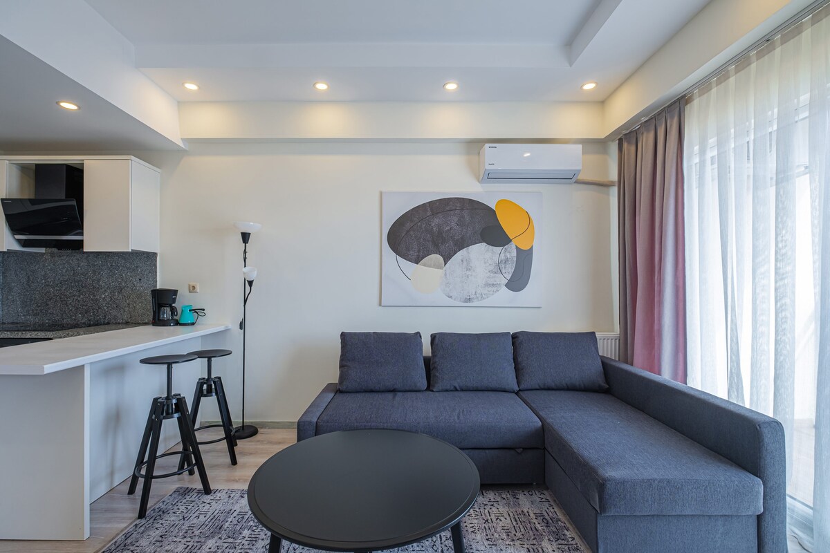 A modern living area features a dark sectional sofa with an abstract art piece on the wall. A round coffee table sits in front, and two black stools are positioned at the nearby kitchen counter. Natural light filters through sheer curtains, enhancing the space.