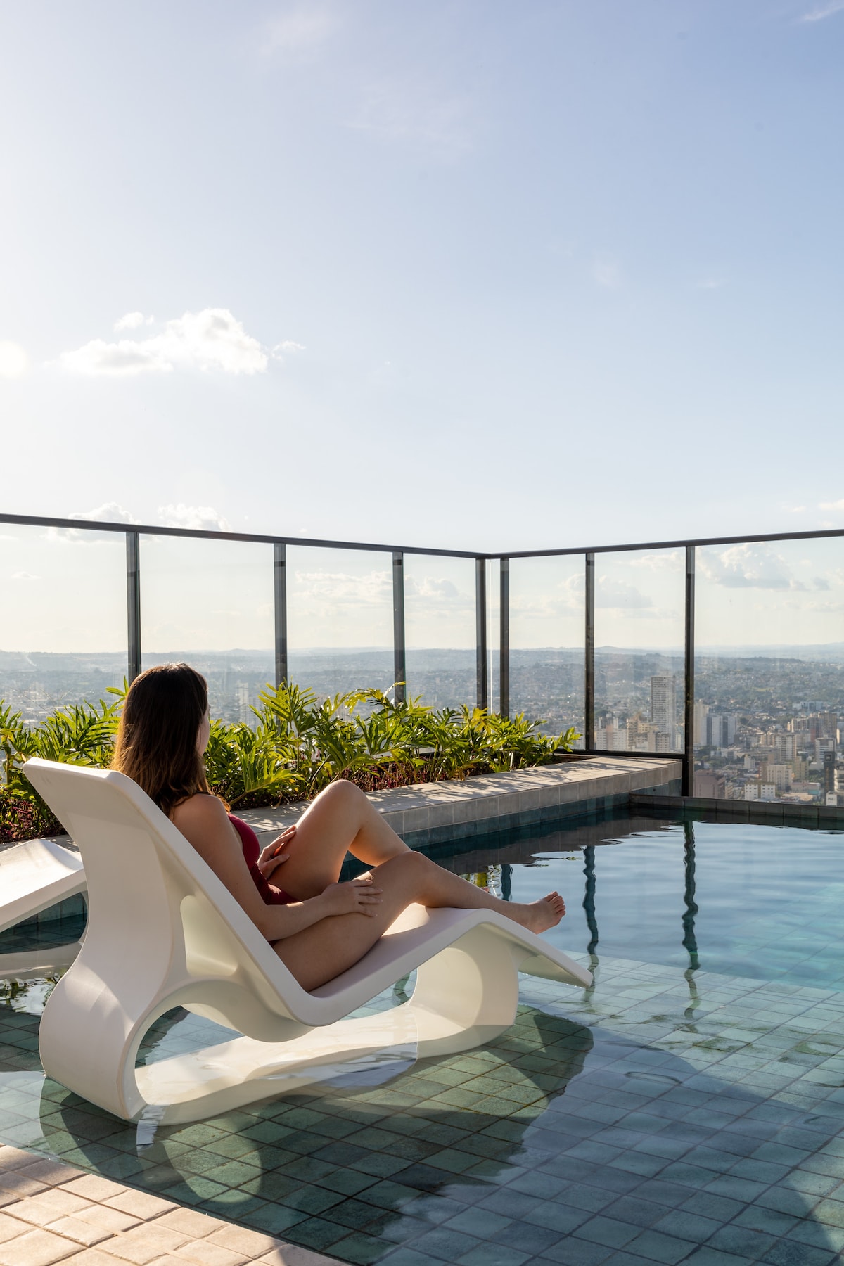 A white lounge chair is positioned at the edge of an infinity pool, overlooking a panoramic view of the city. Lush greenery lines the pool area, with clear glass railings providing an unobstructed sight of the skyline against a bright blue sky.