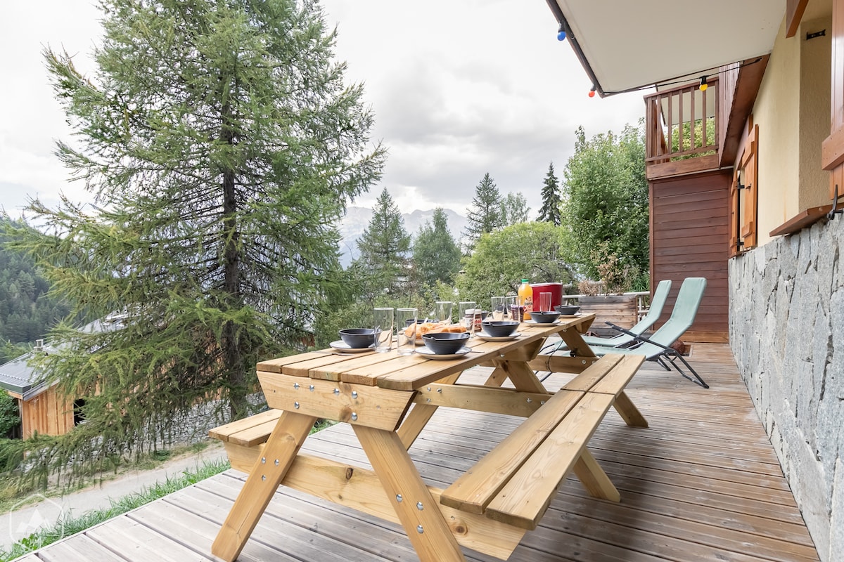 A spacious wooden terrace features a large picnic table set for a meal, with bowls and drinks arranged attractively. Two lounge chairs are positioned beside the table, and the backdrop includes trees and mountain views, creating a relaxed outdoor setting.