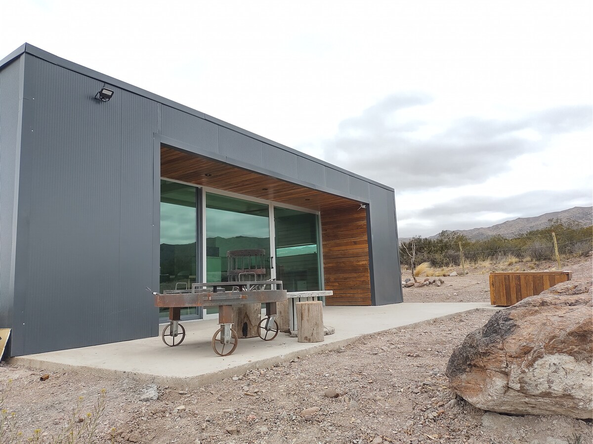 The modern exterior of the house features large glass windows framed by a sleek metal facade. A wooden overhang adds a natural touch. A small outdoor seating area with rustic wooden tables is visible, complemented by the surrounding rocky landscape and distant mountains.