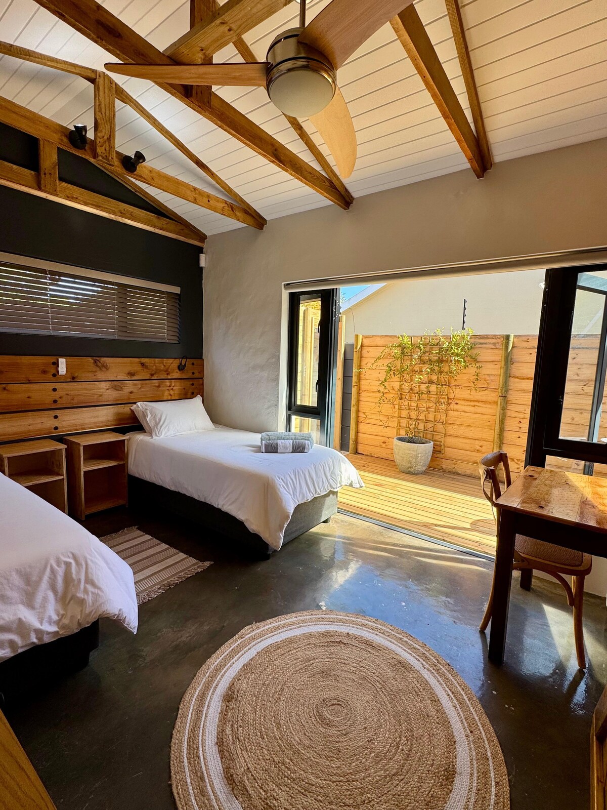 A bright room features two single beds with dark bed bases, positioned beside wooden nightstands. Large windows provide natural light and offer views of a private patio with greenery. A simple desk and rug complement the space, along with a ceiling fan and slanted wooden ceiling.