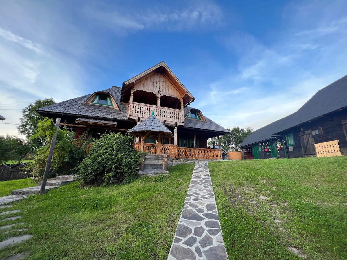 The exterior of the property showcases a wooden house with intricate architectural details, set against a clear blue sky. A stone path leads to the main entrance, surrounded by well-maintained green grass and shrubs.