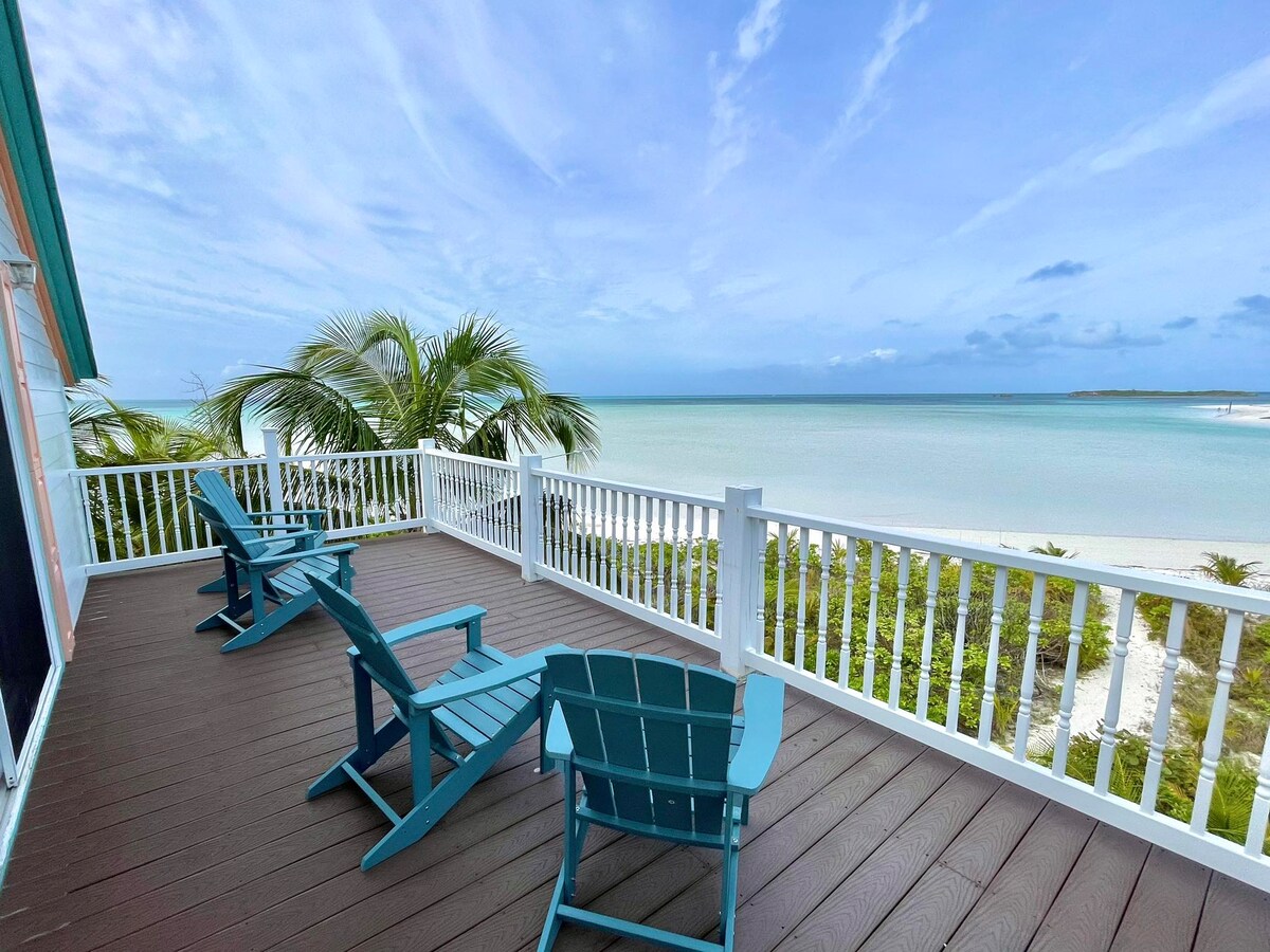 A spacious beachfront balcony features multiple turquoise chairs arranged for relaxation, overlooking a pristine sandy beach and calm turquoise waters. Lush palm trees can be seen nearby, completing the serene coastal setting under a bright sky.