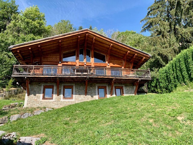 The modern chalet is positioned on a gently sloping grassy area, featuring a wooden exterior with large windows and a spacious balcony. Surrounded by lush greenery, the structure showcases an inviting alpine architecture under a clear blue sky.