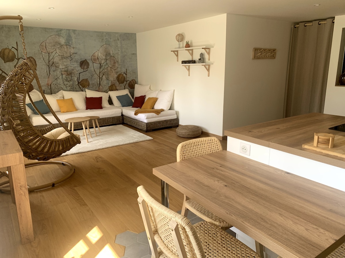 An open living space features a cozy seating area with a large sectional sofa adorned with various cushions. A hanging chair adds a unique touch. A wooden dining table with woven chairs is positioned nearby, complemented by a light and airy ambiance from natural lighting.