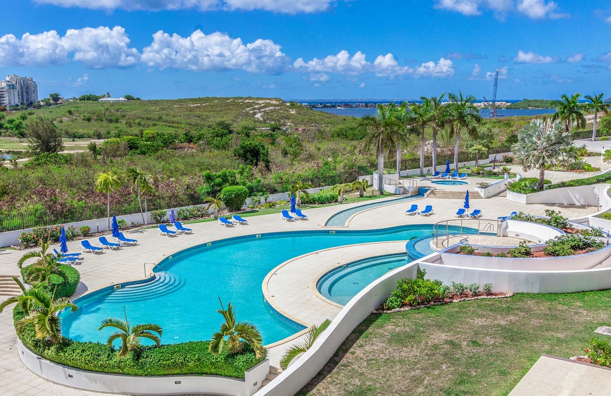 A spacious outdoor pool area is surrounded by lush greenery and palm trees. Multiple lounge chairs are arranged around the pool, which features a unique, flowing design. The tranquil water reflects the clear blue sky and distant coastal landscape, inviting relaxation and enjoyment.