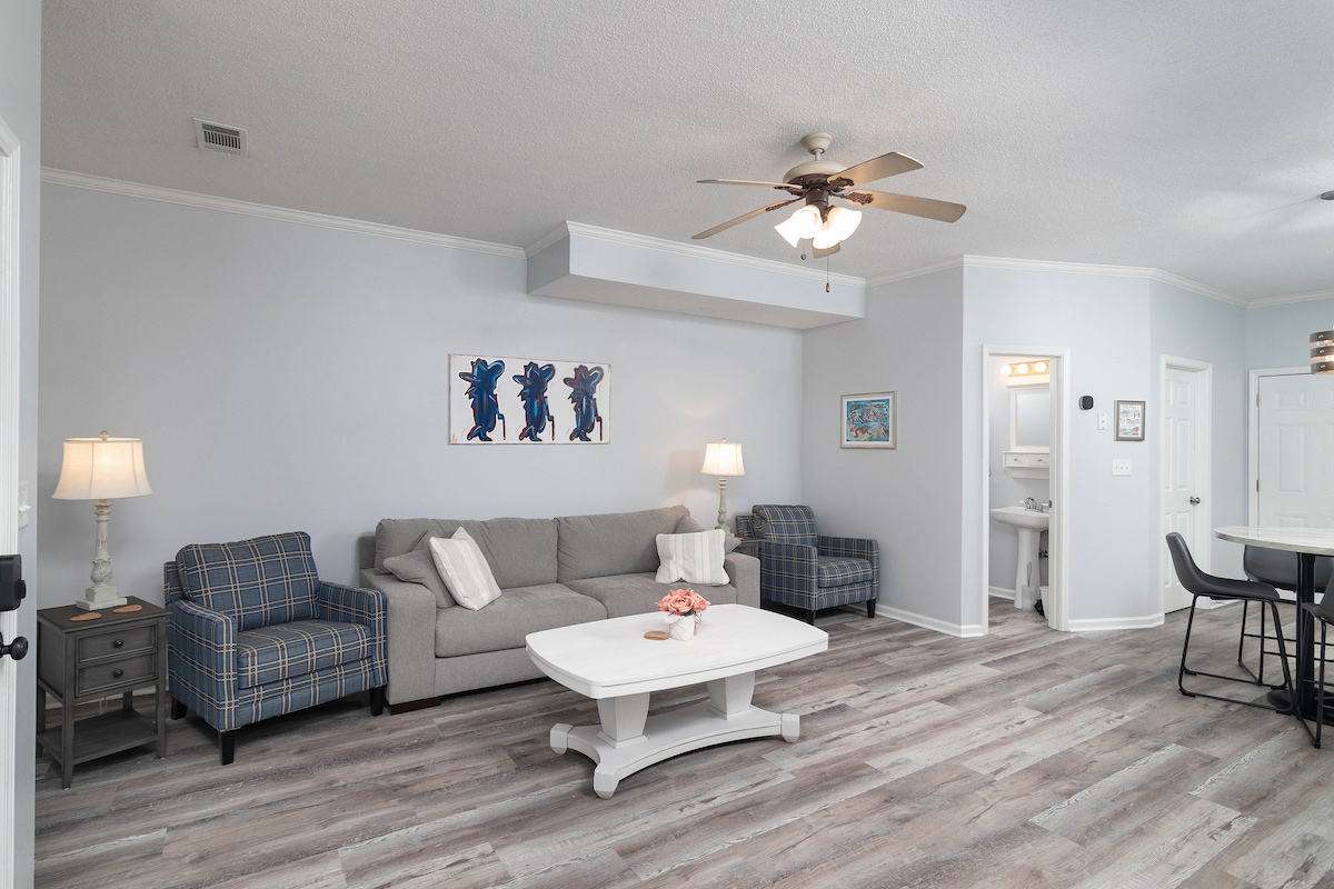 A comfortable living area features a gray sofa and two patterned chairs, accented by a white coffee table. A ceiling fan is present, and artwork adorns the wall. The flooring is light-colored and wood-look, providing a cozy atmosphere.