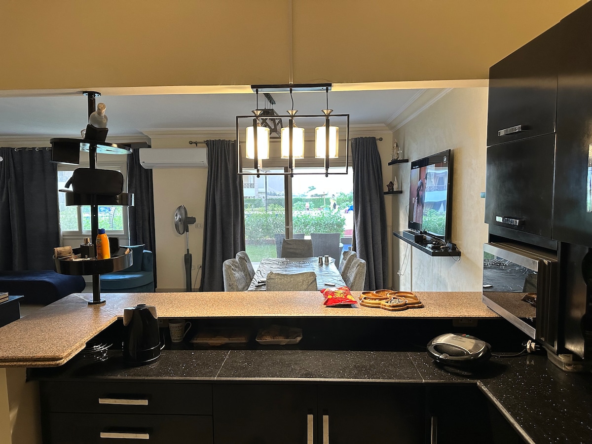 A modern kitchen is featured with sleek black cabinetry and a countertop that overlooks the dining area. The dining table accommodates six and is set beside large windows with grey curtains. A stylish lighting fixture hangs above the table, and a television is mounted on the wall.