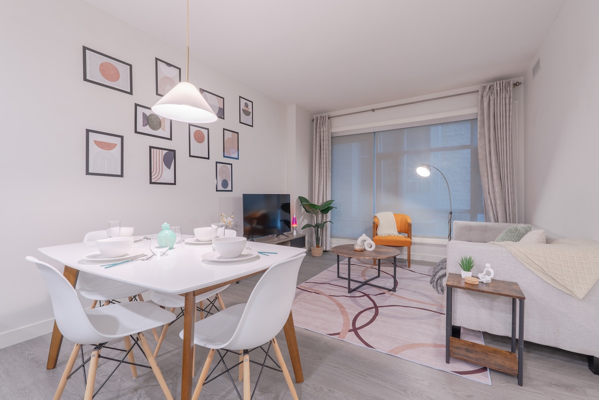 An open living area is presented with a modern dining table, six white chairs, and a cozy seating arrangement featuring a light gray couch and an orange accent chair. A TV is positioned beside a large window dressed with light curtains, allowing natural light to fill the space.