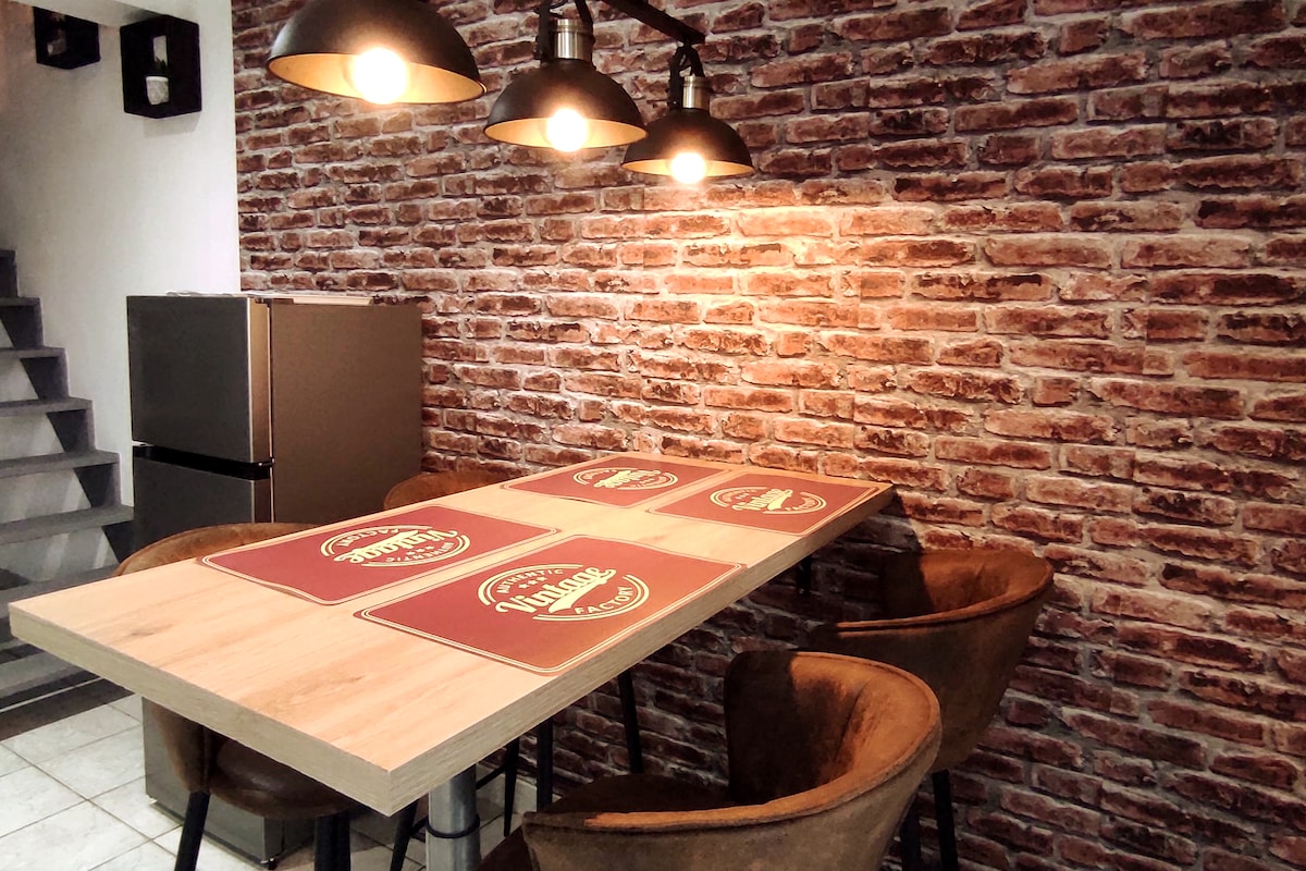 A dining area features a wooden table with decorative placemats, surrounded by four upholstered chairs. A textured brick wall adds character, while three pendant lights offer warm illumination above the table. The space is complemented by a glimpse of a modern refrigerator in the background.