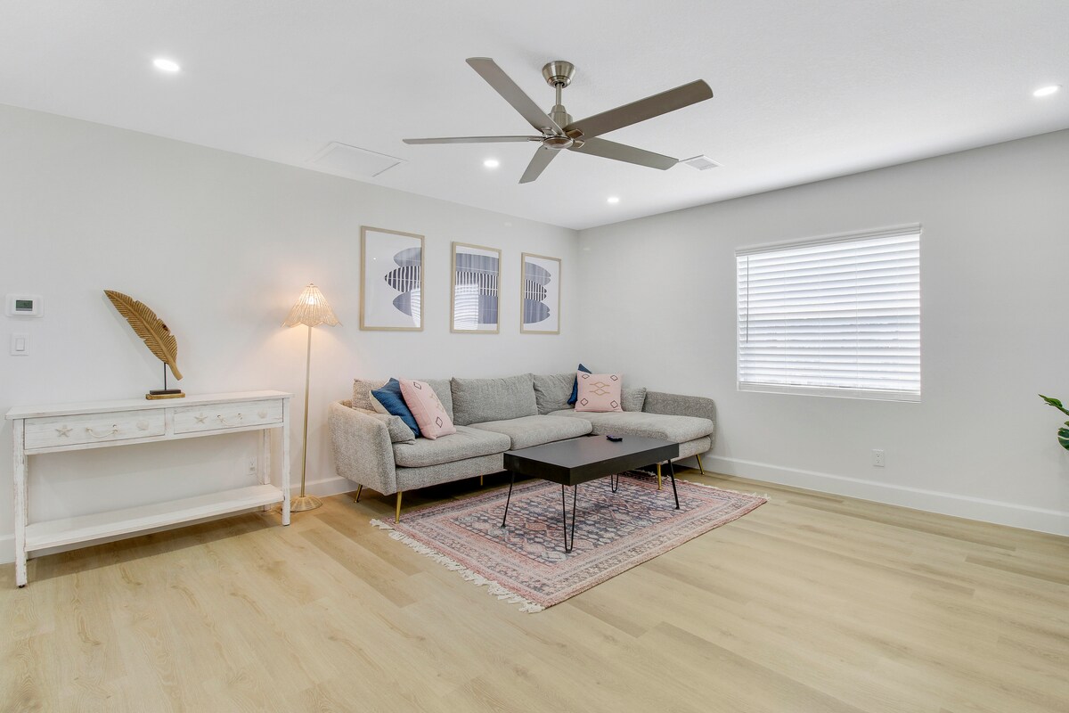 A light-filled living area features a modern sectional sofa arranged around a low coffee table. Three framed artworks are displayed on the wall above, and a stylish console table is positioned to the left. Natural wood flooring adds warmth to the space.