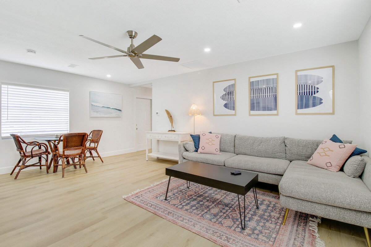 A spacious living area features a light grey sectional couch, a sleek black coffee table, and a patterned area rug. Natural light enters through the window beside a wooden dining table with four rattan chairs, enhancing the room's modern and comfortable ambiance.