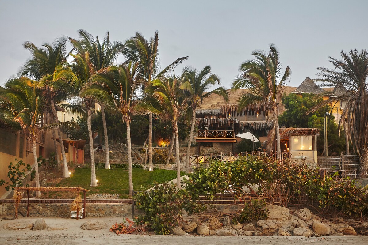 The exterior of the house showcases a lush landscape with palm trees lining the property. The vibrant greenery is complemented by stone paths and rustic wooden structures. Lighting illuminates the area, creating a calm ambiance as the beach and distant hills set a serene backdrop.