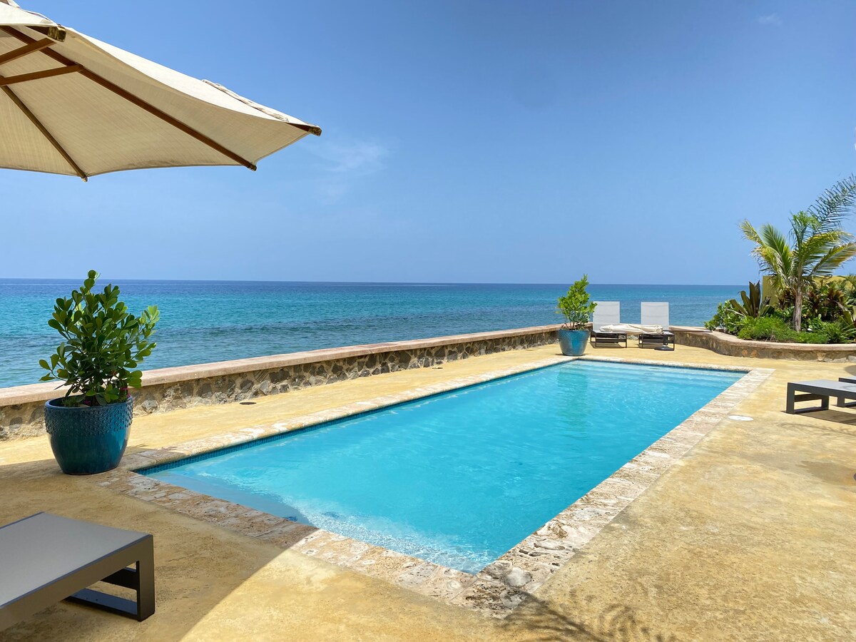 A serene beachfront pool is surrounded by a stone wall and lush greenery. Two lounge chairs are positioned nearby, complemented by decorative potted plants. The clear blue water of the pool reflects the vibrant hues of the ocean, enhancing the relaxing atmosphere.