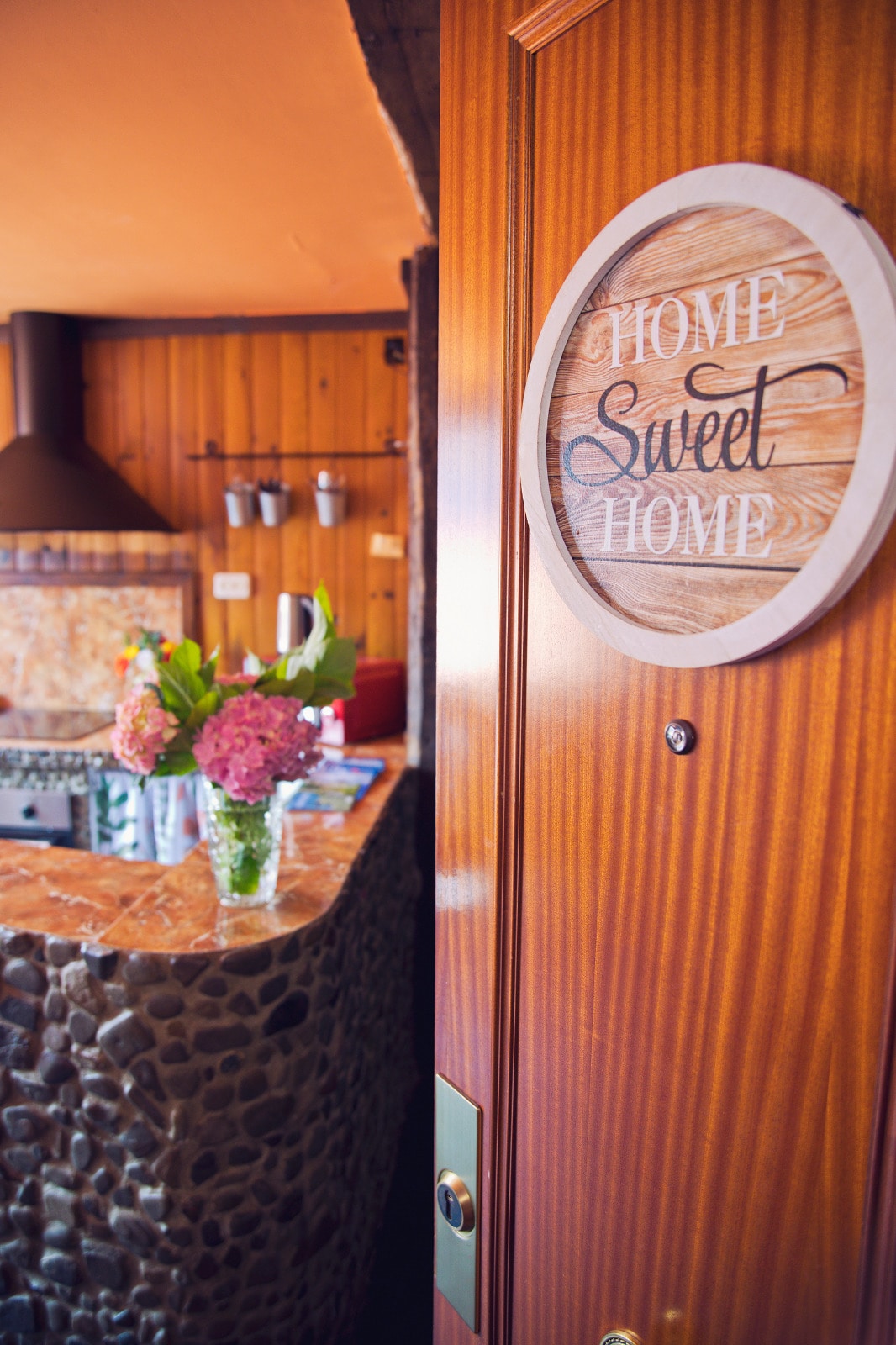 A wooden door is framed by a warm-toned entrance, featuring a decorative sign that reads 'Home Sweet Home.' A welcoming atmosphere is presented by a kitchen area in the background, where fresh flowers are arranged in a vase on a stone counter.