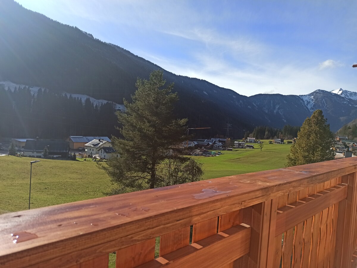 A wooden balcony railing offers an expansive view of the mountainous landscape and green fields below. Tall trees frame the scene, with distant houses dotting the valley and a clear blue sky above, suggesting a serene atmosphere.