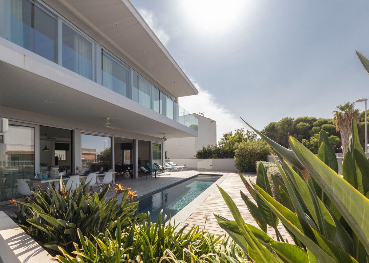 A modern villa is showcased, featuring large glass windows that reflect natural light. An inviting saltwater pool lies adjacent to a wooden deck, surrounded by lush greenery. The outdoor area offers space for relaxation, with ample seating available for guests.