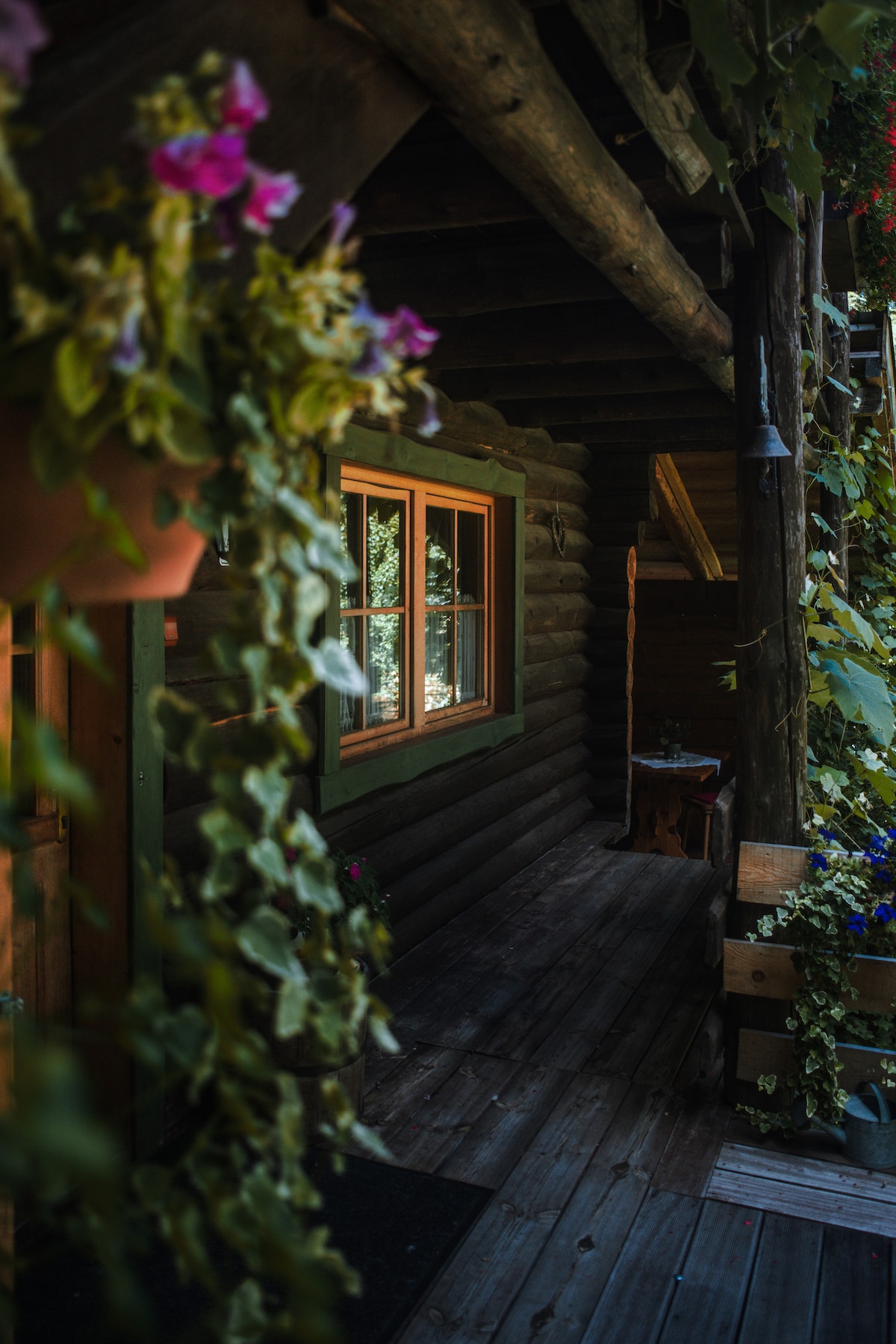 A wooden entryway is framed by lush greenery, with flower pots hanging alongside the door. A warm light emanates from the window, inviting guests to enter. The rustic wooden beams and flooring convey a natural setting.