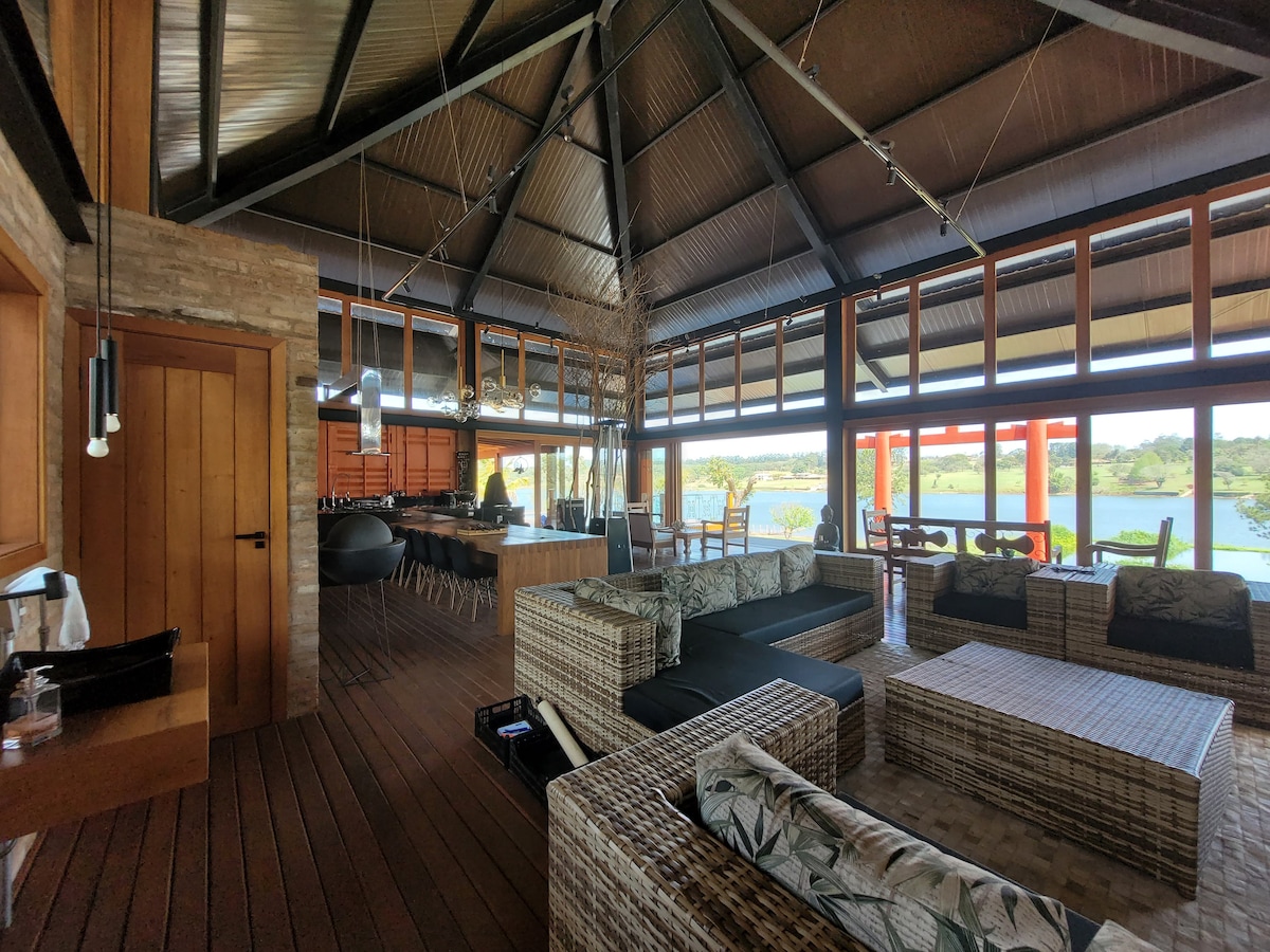 An expansive lounge area is characterized by high ceilings and large windows, providing natural light and views of the surrounding lake. Comfortable seating arrangements made of woven materials are positioned around a central coffee table, creating an inviting space for relaxation and socializing.