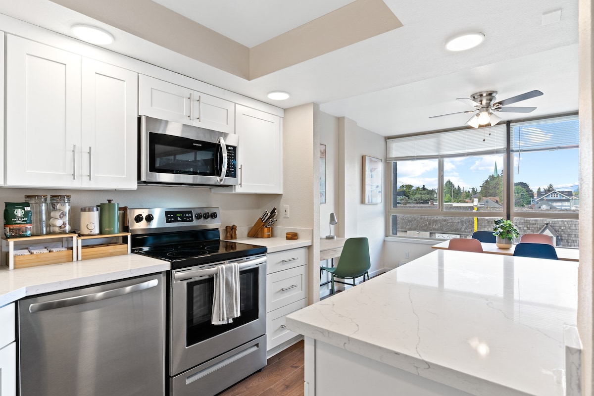 A modern kitchen is seen with white cabinetry and stainless steel appliances. A marble island provides additional workspace, while a dining table with colorful chairs is positioned nearby. Large windows allow natural light to brighten the space, offering a view of the outside.