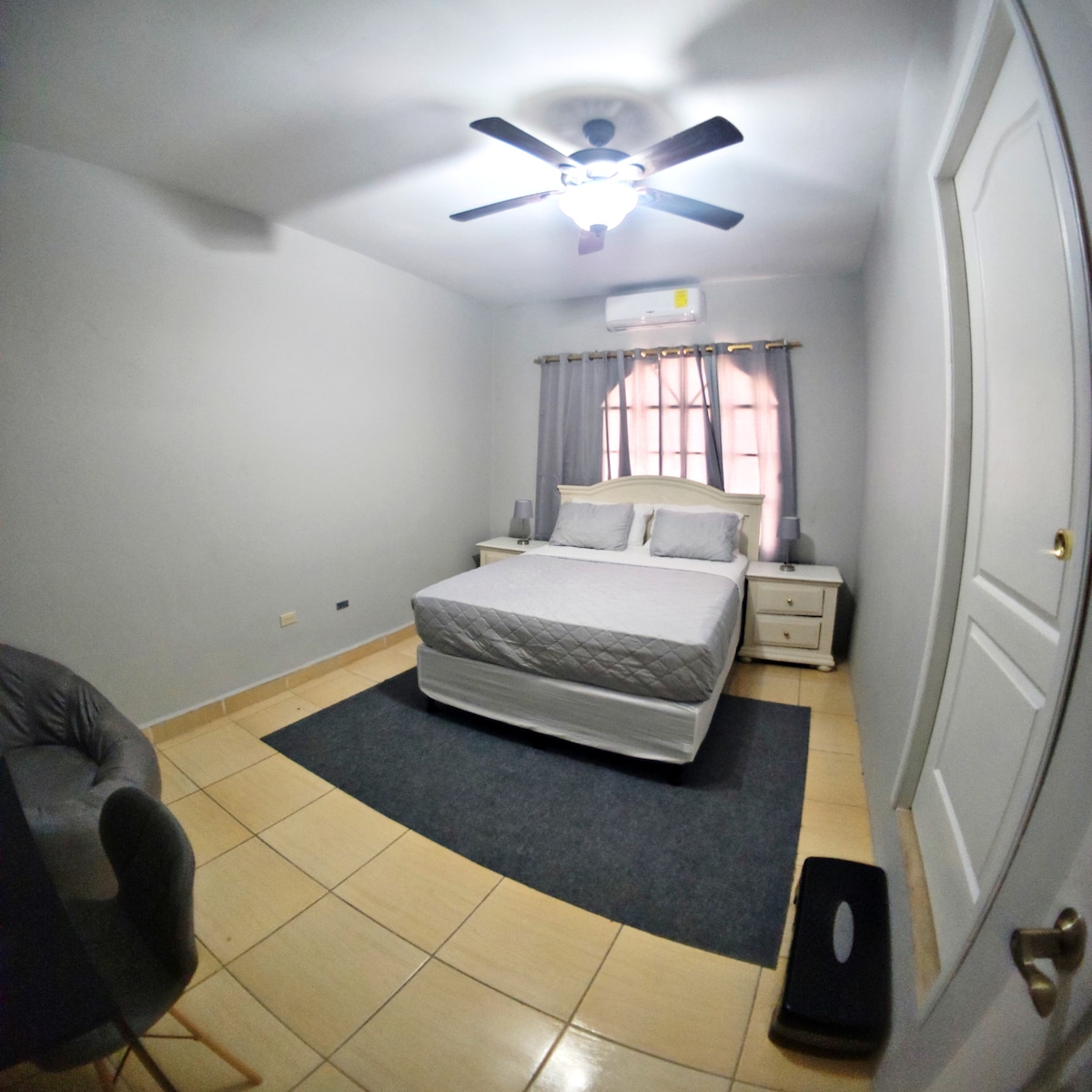 A well-lit bedroom features a spacious bed with gray linens. Two bedside tables are positioned beside the bed, and a fan is mounted on the ceiling. Natural light enters from a window adorned with sheer curtains. A comfortable chair and a small rug complete the space.