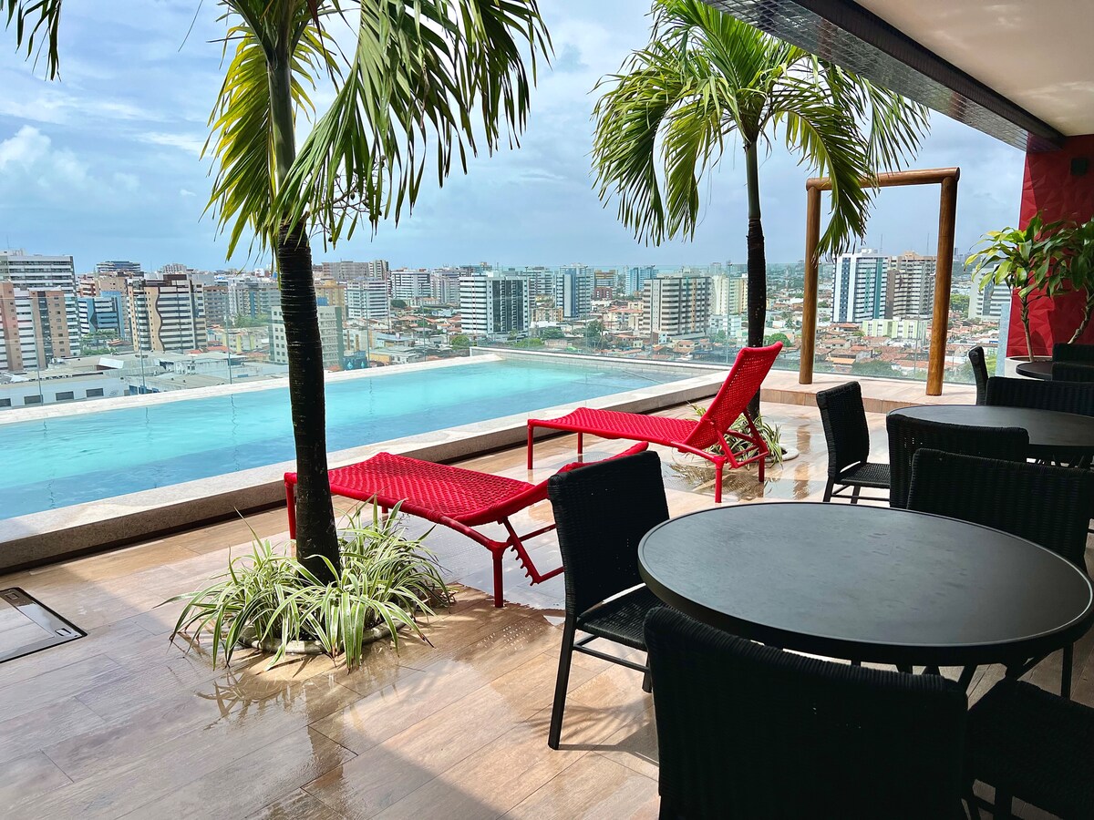 An outdoor area is presented with lounge chairs by a glass-panel infinity pool. Lush palm trees frame the view of the city skyline under a cloudy sky. Black dining furniture is placed nearby, inviting relaxation and enjoyment of the scenery.